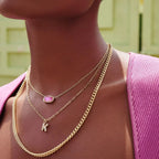 Person wearing gold necklaces with a pink gemstone and a small pendant against a blurred background.