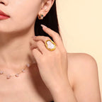 Close-up of a woman wearing gold earrings and a gold ring with a large gemstone on a beige background