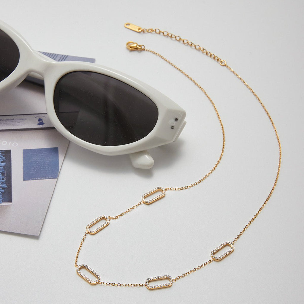 Gold chain necklace on a light gray surface with white sunglasses and a magazine.