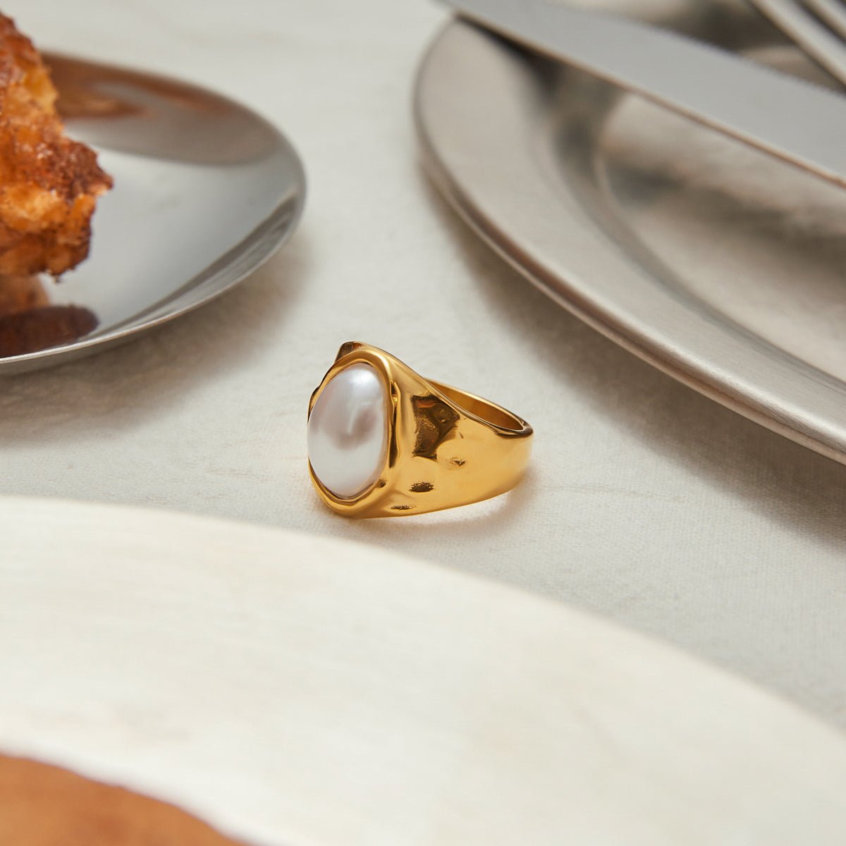 Gold ring with a pearl on a table setting with plates and cutlery.