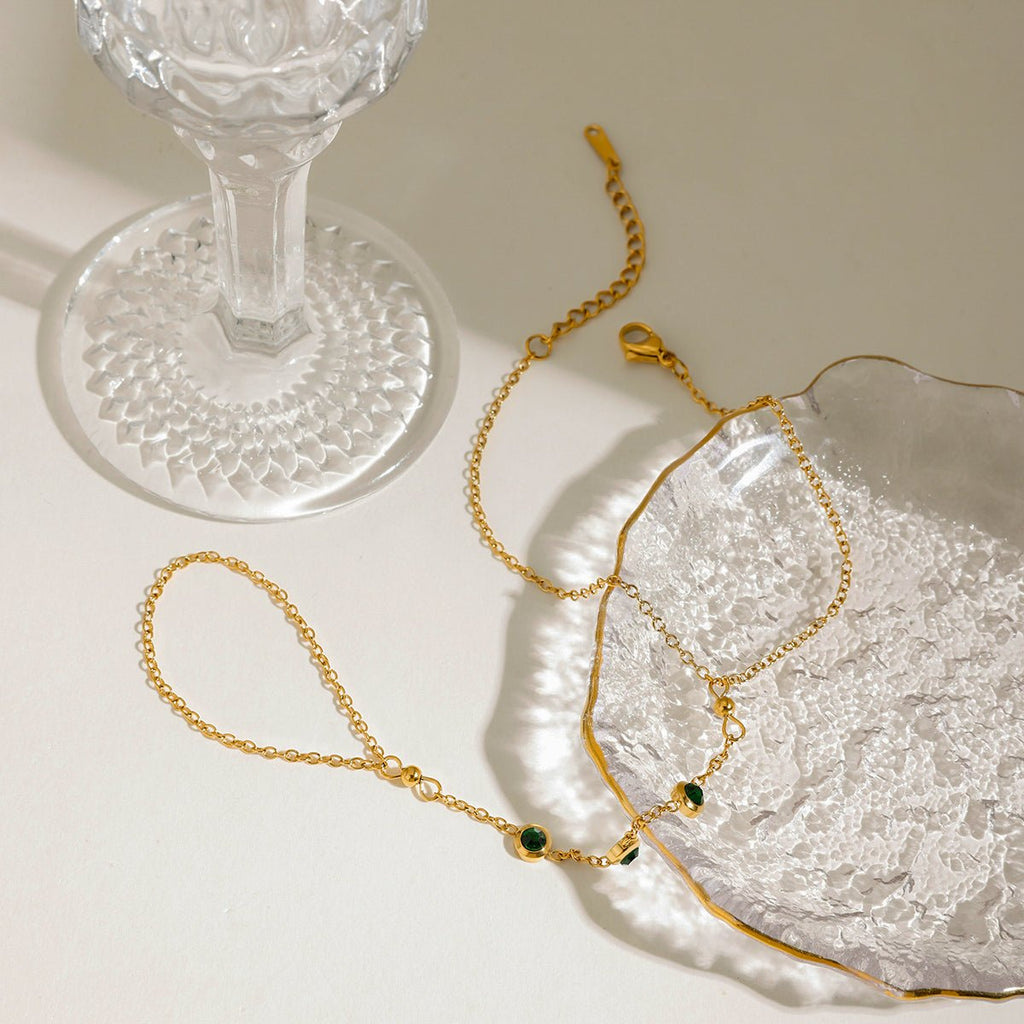 Gold bracelet with green stones on a textured white surface next to a clear glass object.