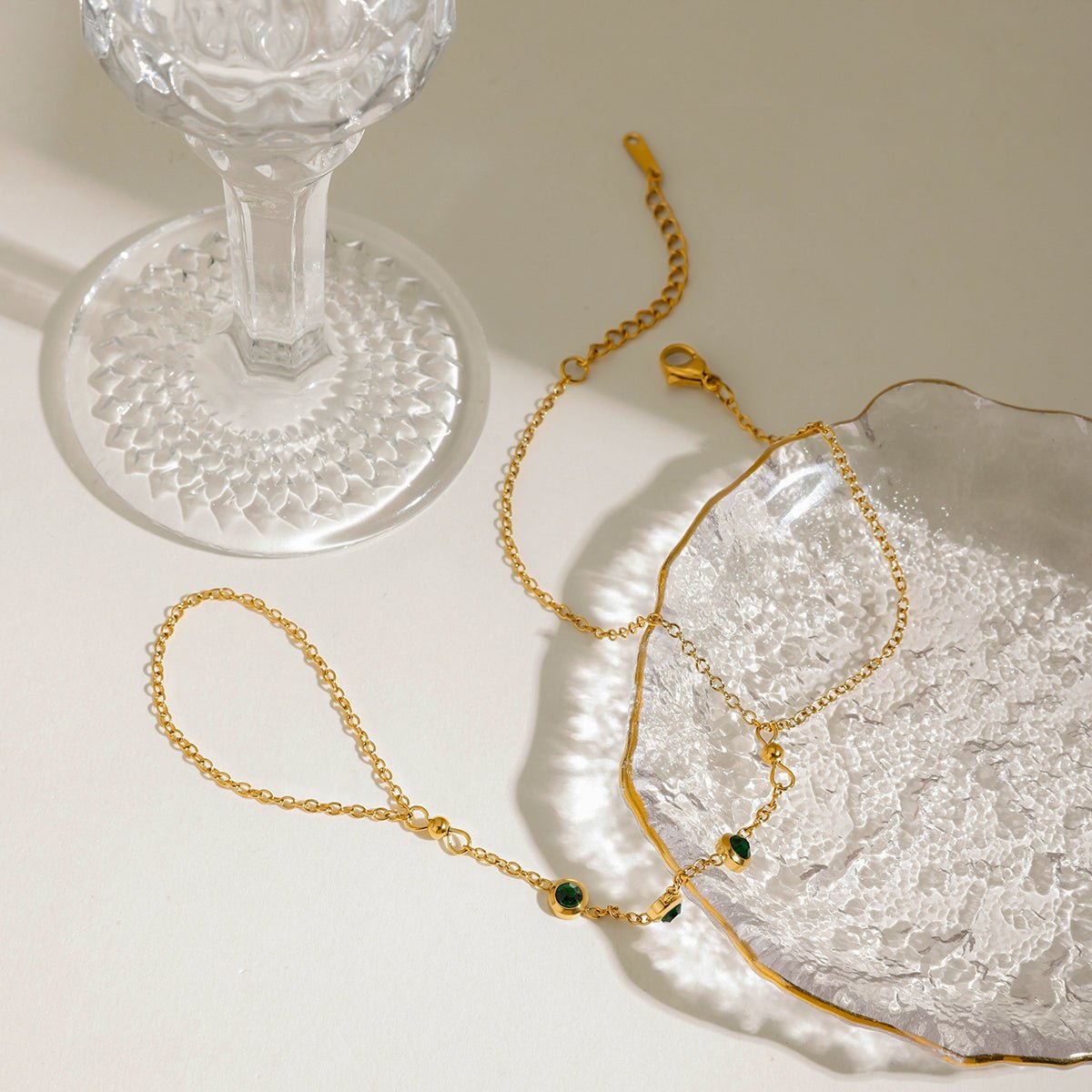 Gold bracelet with green stones on a textured white surface next to a clear glass object.