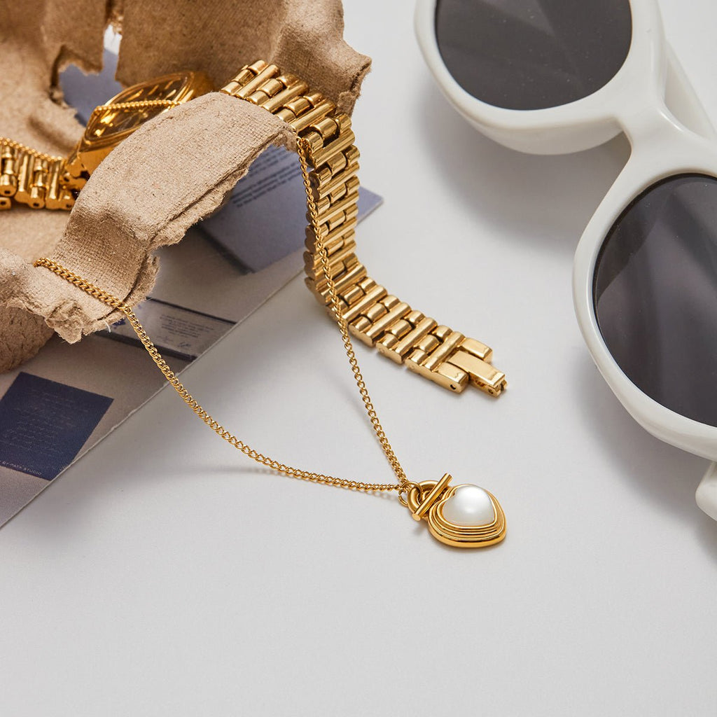 Gold bracelet with a heart-shaped pendant on a light surface next to sunglasses and a cup.