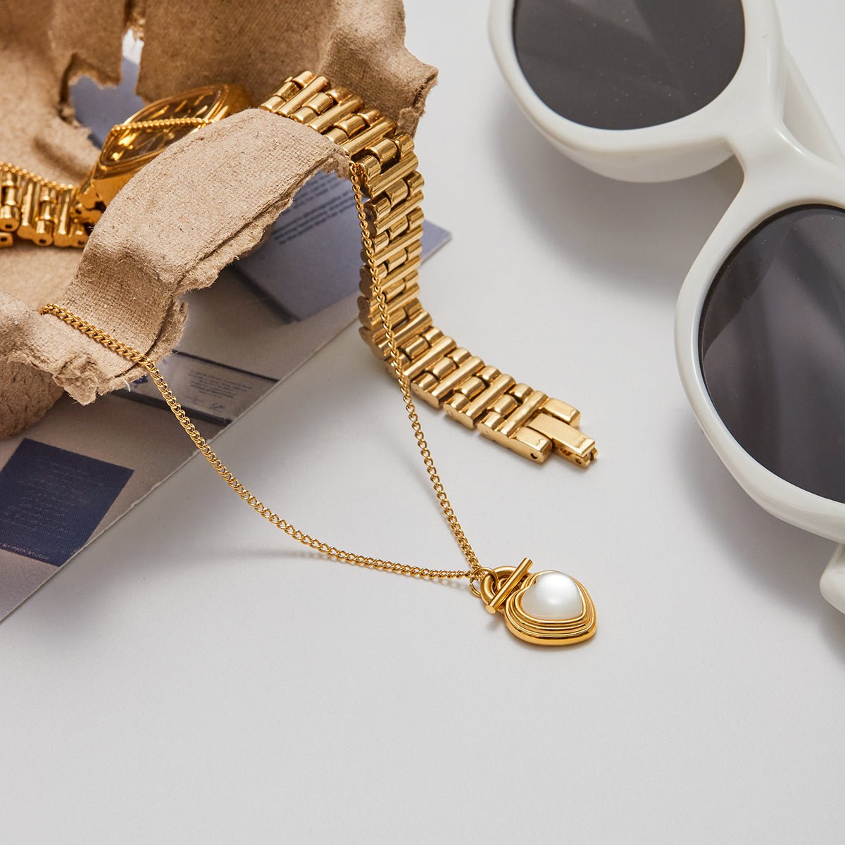 Gold bracelet with a heart-shaped pendant on a light surface next to sunglasses and a cup.