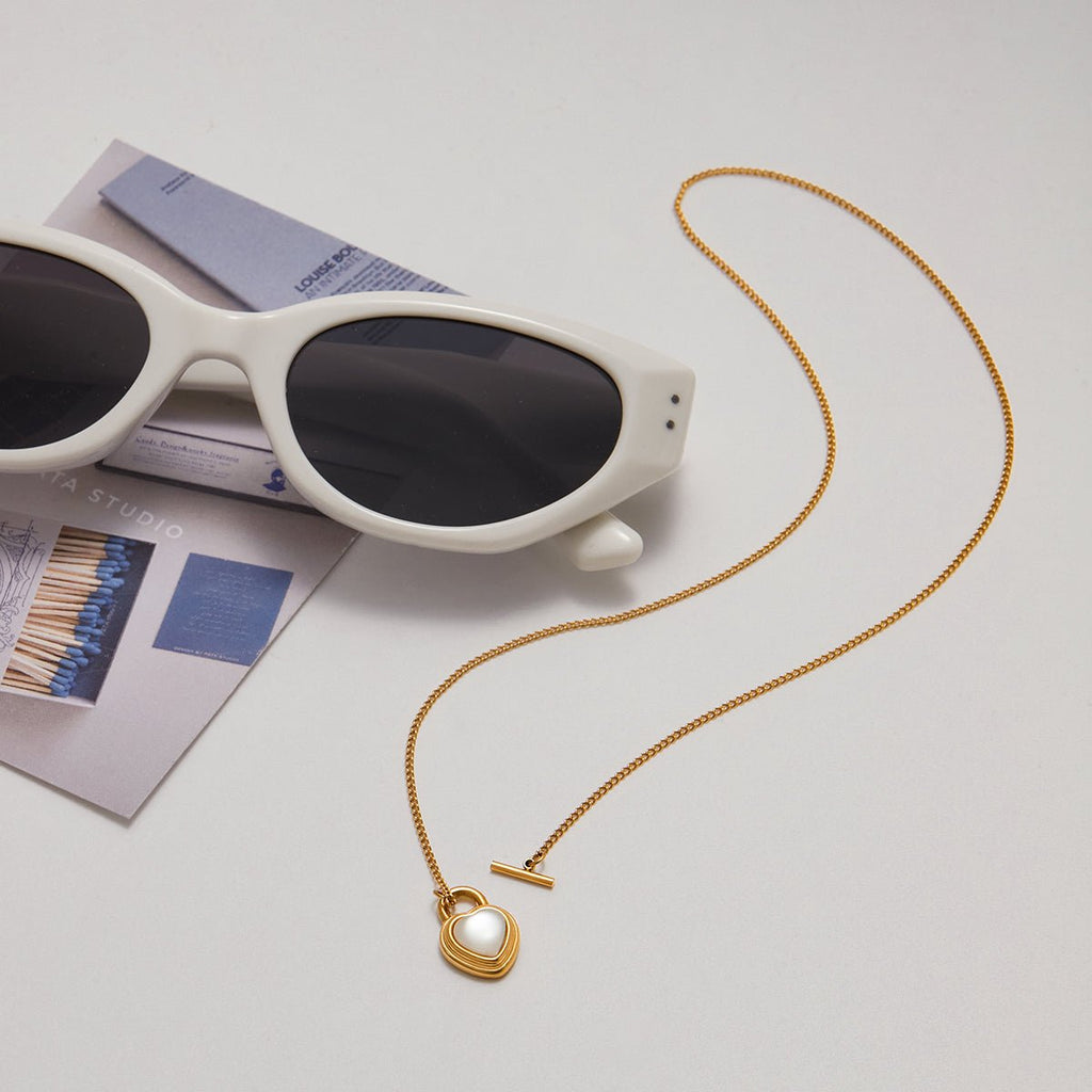 Gold necklace with a heart pendant next to white sunglasses on a light gray surface.