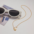 Gold necklace with a heart pendant next to white sunglasses on a light gray surface.