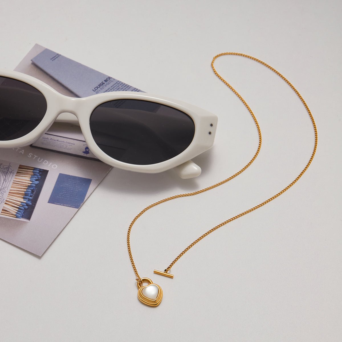 Gold necklace with a heart pendant next to white sunglasses on a light gray surface.