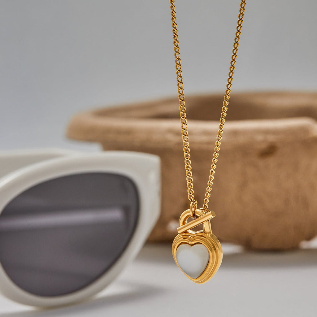 Gold necklace with a heart-shaped pendant next to sunglasses on a neutral background
