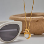 Gold necklace with a heart-shaped pendant next to sunglasses on a neutral background