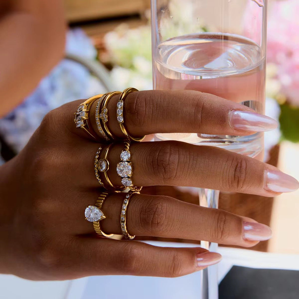 Hand wearing multiple gold rings with gemstones holding a glass of water.