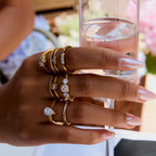 Hand wearing multiple gold rings with gemstones holding a glass of water.