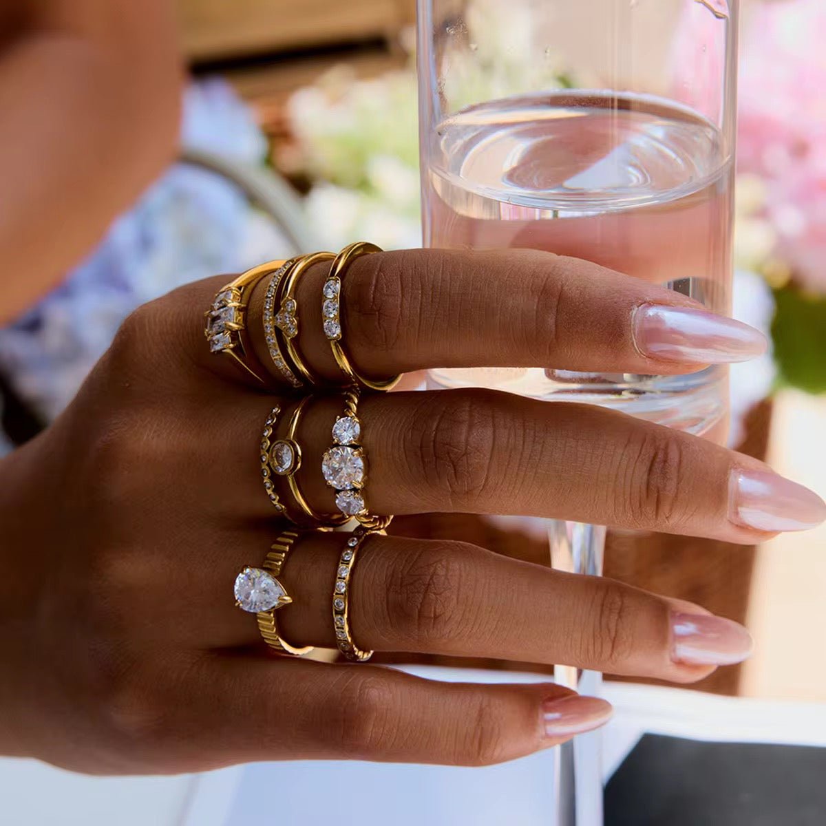 Hand wearing multiple gold rings with gemstones holding a glass of water.