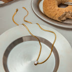 Gold chain necklace with a heart pendant on a white plate, with a bagel in the background.