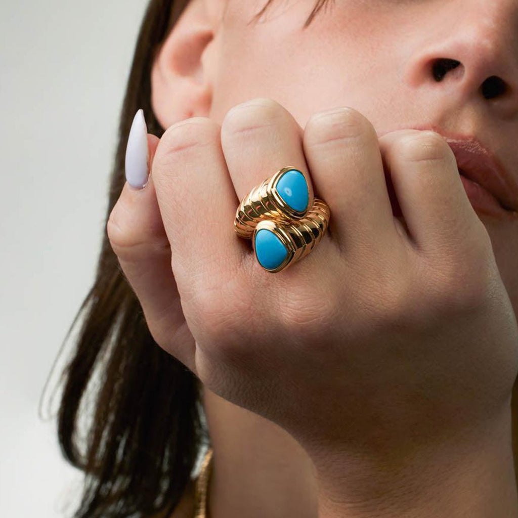 Close-up of a hand wearing a gold ring with turquoise stones against a neutral background