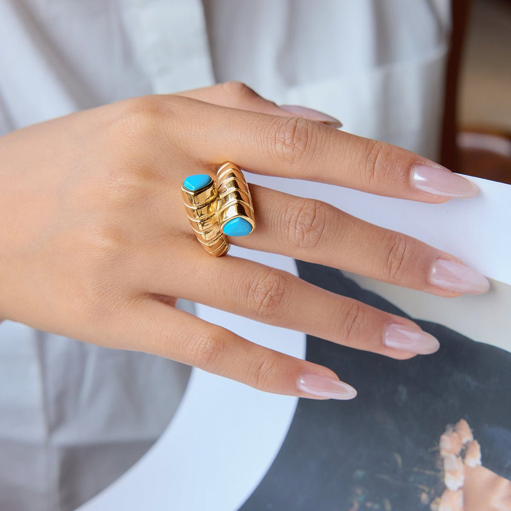 Hand wearing a gold ring with blue stones on a neutral background