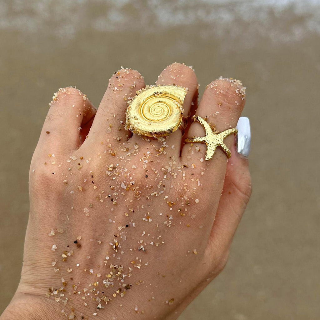 Hand wearing gold shell and starfish rings with sand on a beach.