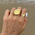 Hand wearing gold shell and starfish rings with sand on a beach.