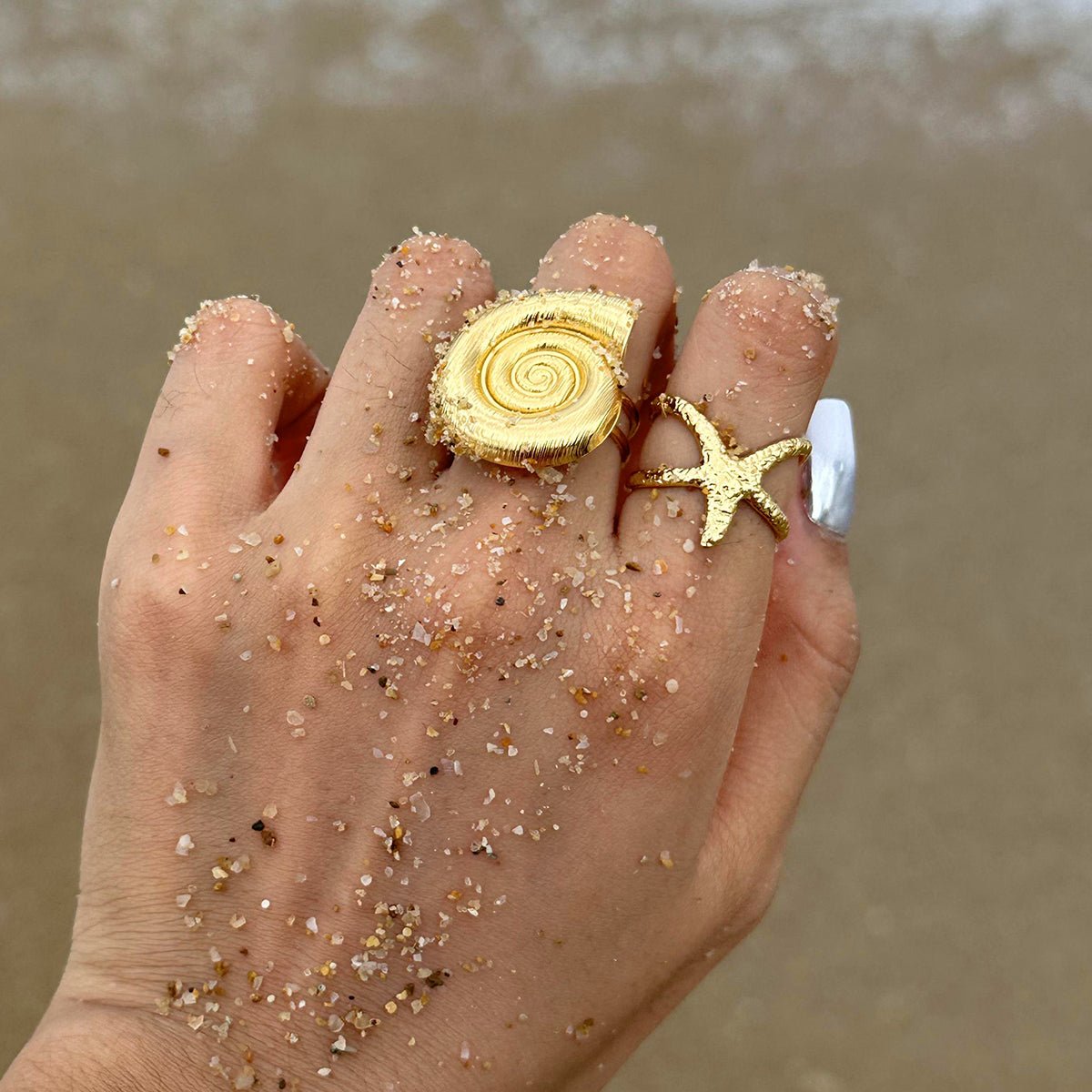 Hand wearing gold shell and starfish rings with sand on a beach.
