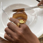 Hand wearing a gold ring with a starfish design, holding a knife and fork over a plate with a croissant.