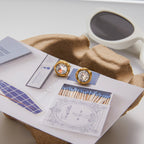 Gold earrings with clear stones on a matchbook next to a cup of coffee.