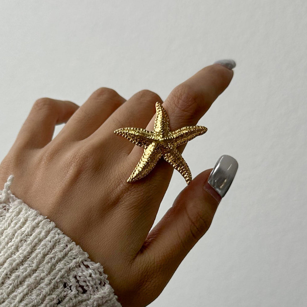 Hand wearing a gold starfish-shaped ring on a neutral background