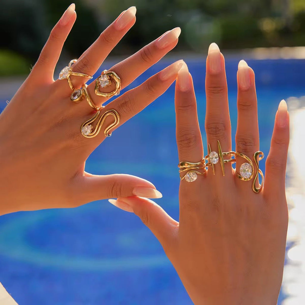 Two hands wearing gold rings with gemstones against a blurred outdoor background