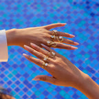Hands wearing multiple gold rings with gemstones against a blue mosaic tile background