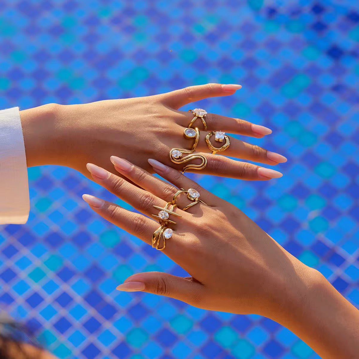 Hands wearing multiple gold rings with gemstones against a blue mosaic tile background