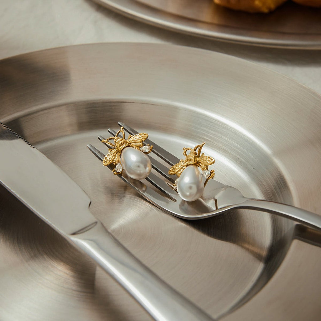 Gold bee and pearl earrings on a fork with a metallic background