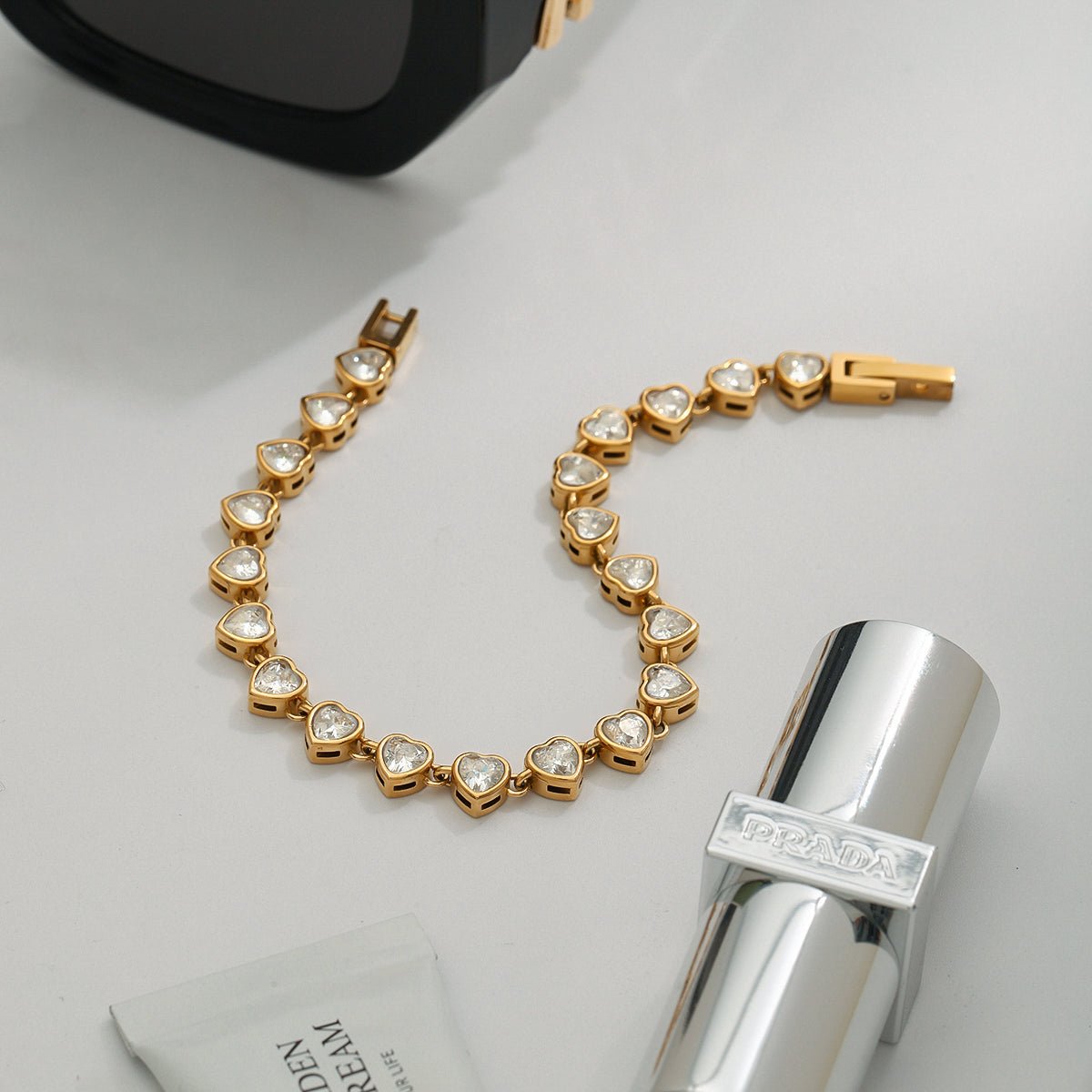 Gold bracelet with heart-shaped stones on a light gray surface with a silver bottle and black object.