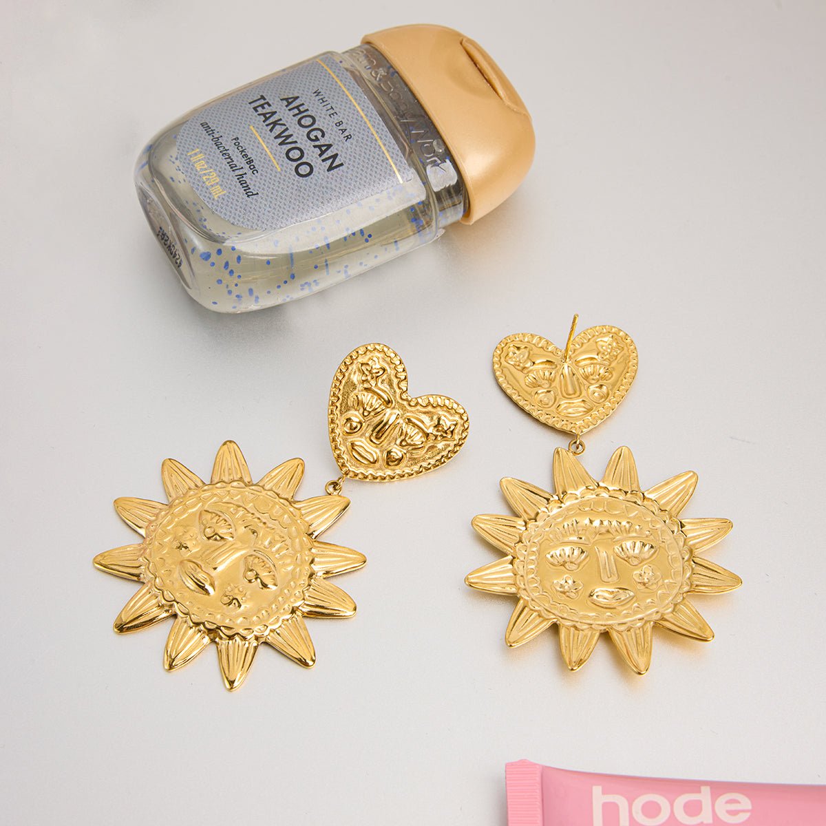 Gold sun and heart-shaped earrings with a bottle of hand sanitizer on a light gray background.
