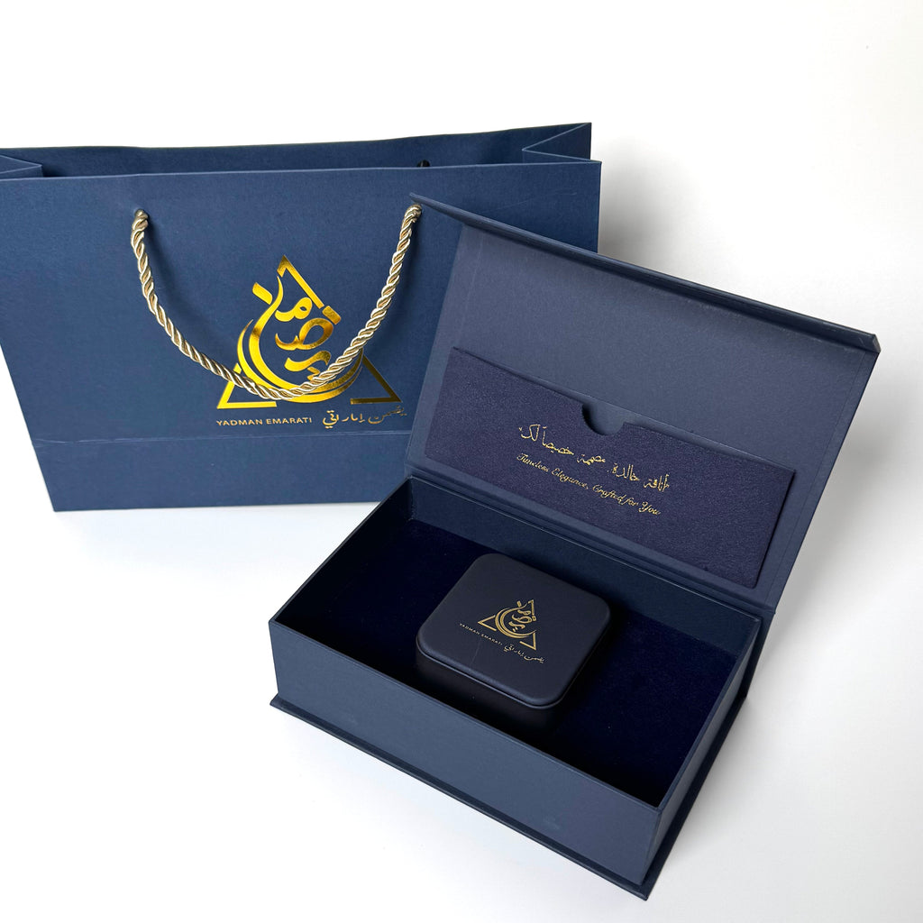 Open navy blue gift box with a black square item inside, accompanied by a matching navy blue bag with gold logo.