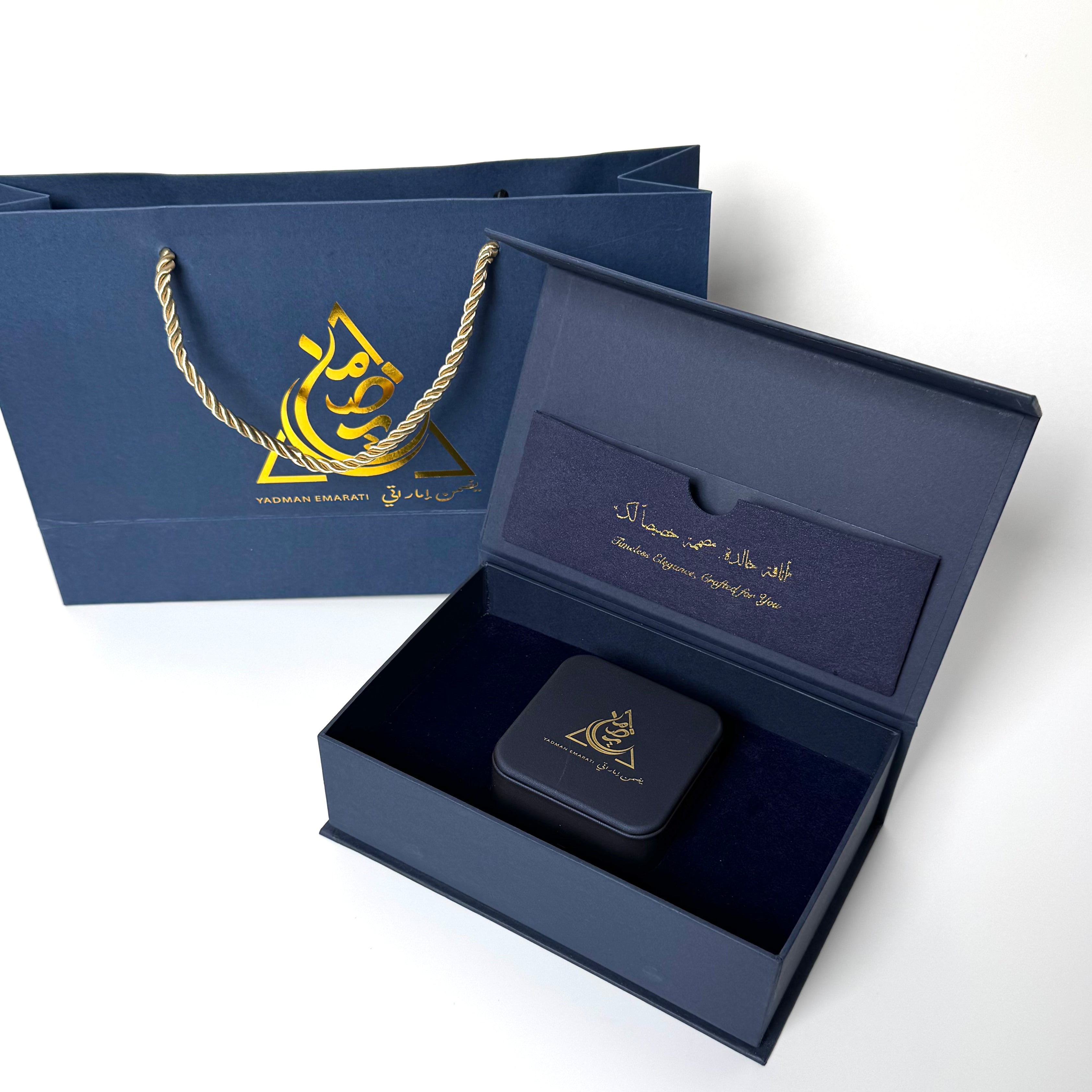 Open navy blue gift box with a black square item inside, accompanied by a matching navy blue bag with gold logo.