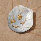 Gold necklaces with sea-themed pendants on a shell against a sandy background