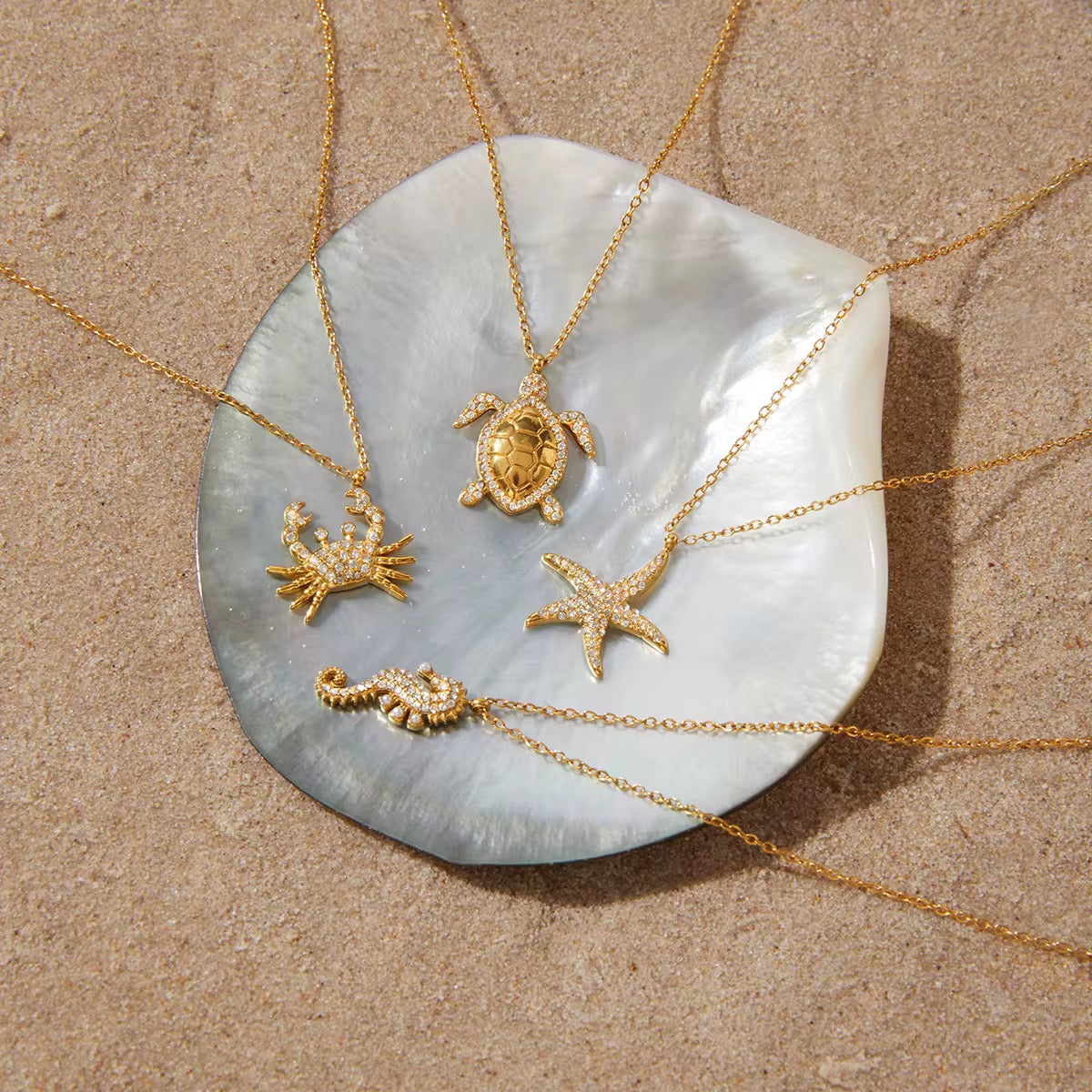 Gold necklaces with sea-themed pendants on a shell against a sandy background