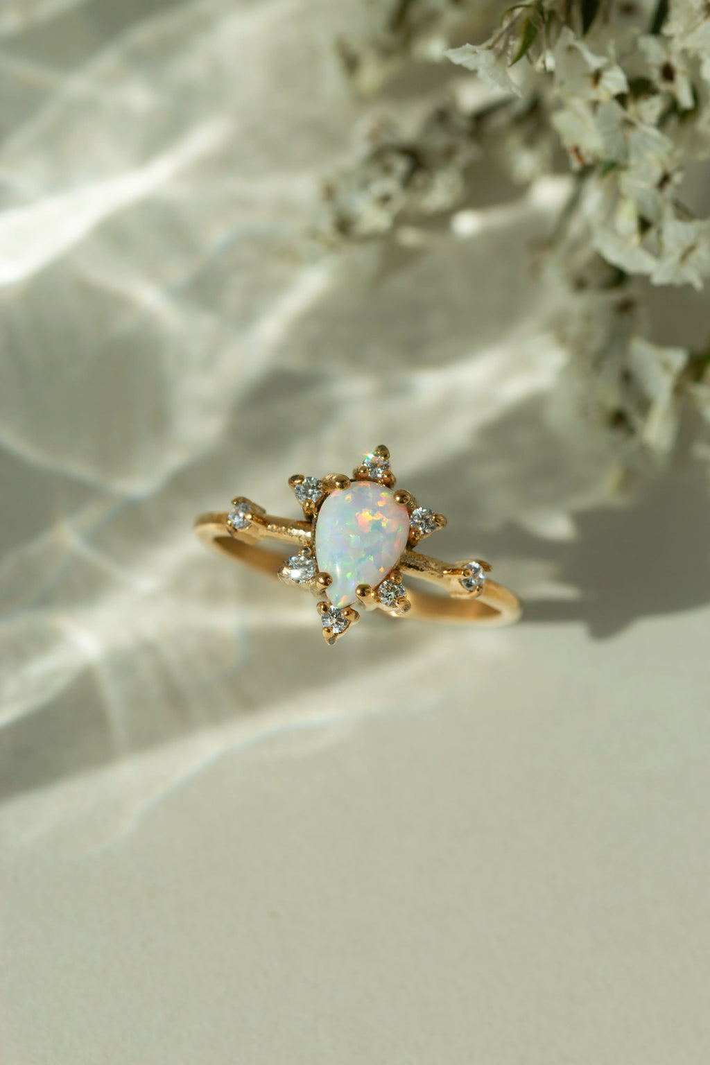 Pear shaped natural opal ring in 14k yellow gold with a diamond cluster starburst halo. Dainty teardrop gemstone engagement ring styled on a cream surface under soft sunlight.