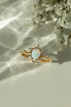 Pear shaped natural opal ring in 14k yellow gold with a diamond cluster starburst halo. Dainty teardrop gemstone engagement ring styled on a cream surface under soft sunlight.