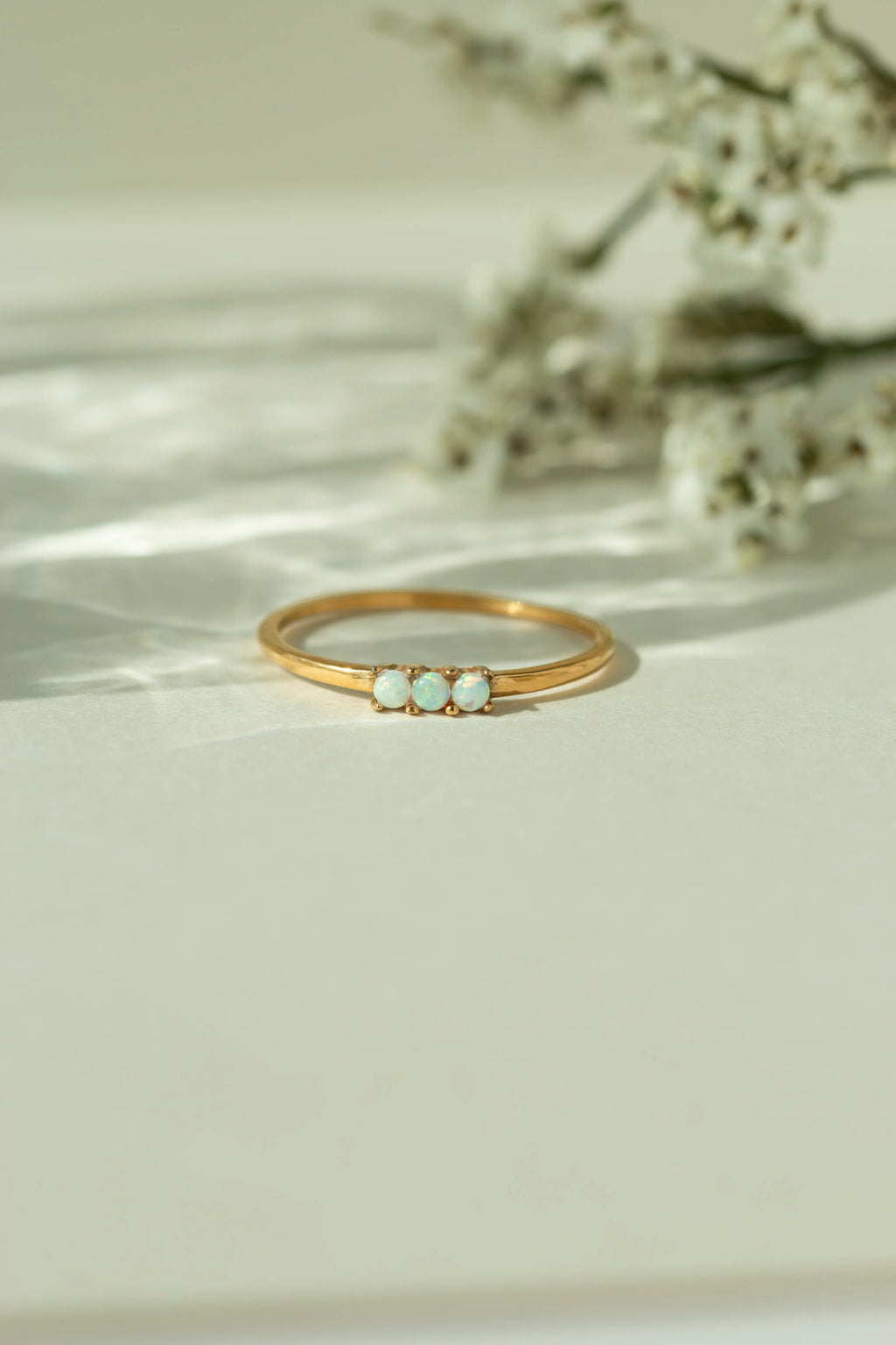 Dainty three stone natural opal ring in a thin 18k yellow gold band. Minimalist triple gemstone stacking ring displayed on a light surface with soft sun shadows.