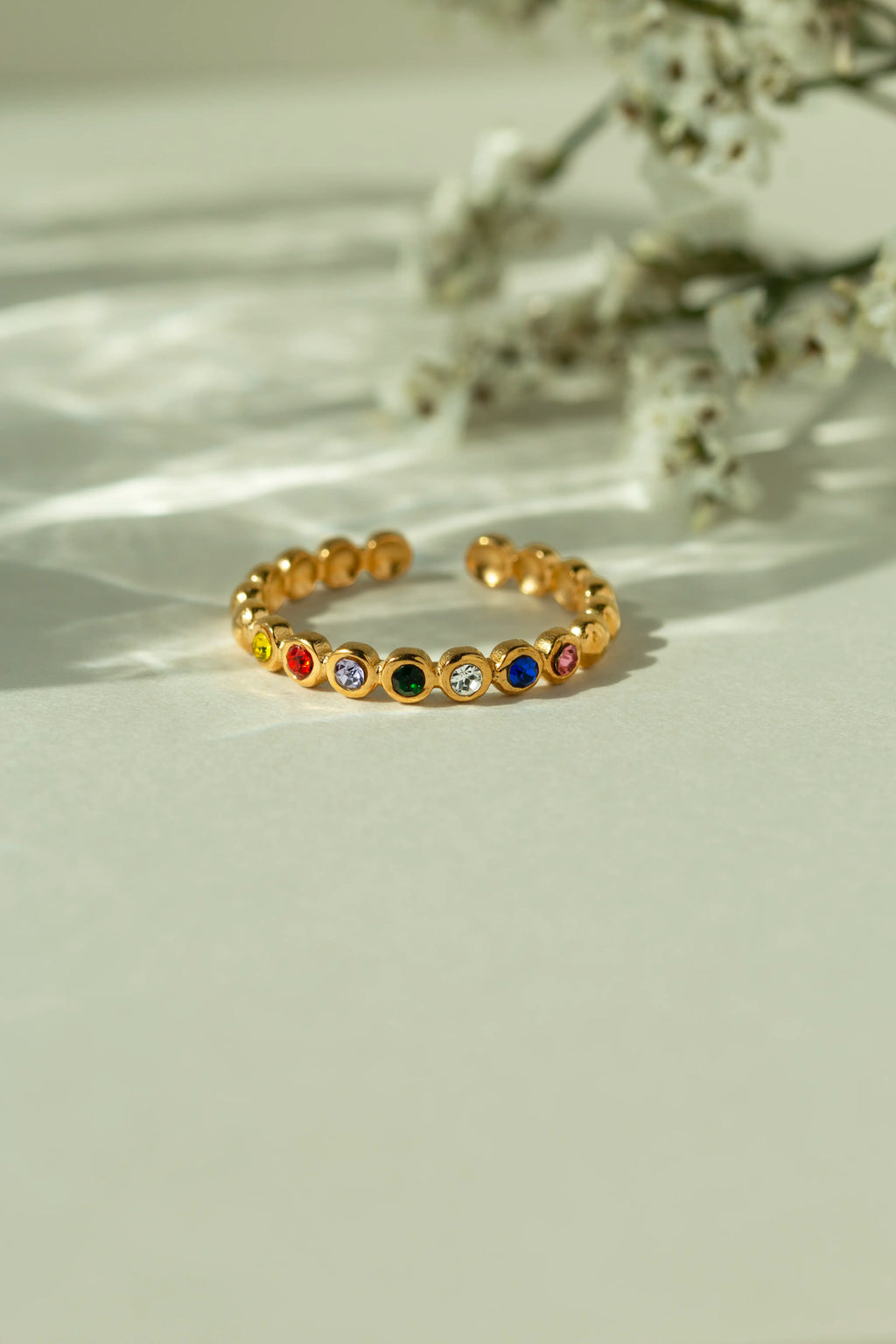  Gold adjustable rainbow bubble ring with round multi color bezel set gemstones. Trendy open back eternity style stacking band displayed on a sunlit neutral background.