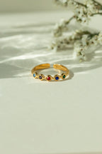 Gold adjustable multi color gemstone ring with colorful birthstones in diamond shaped settings. Trendy open back stackable band displayed on a sunlit neutral background.