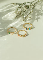 Three 14k yellow gold baguette gemstone rings featuring colorful emerald sapphire and ruby birthstone crystals in scattered cluster settings. Minimalist gold stackable bands displayed on a sunlit background.