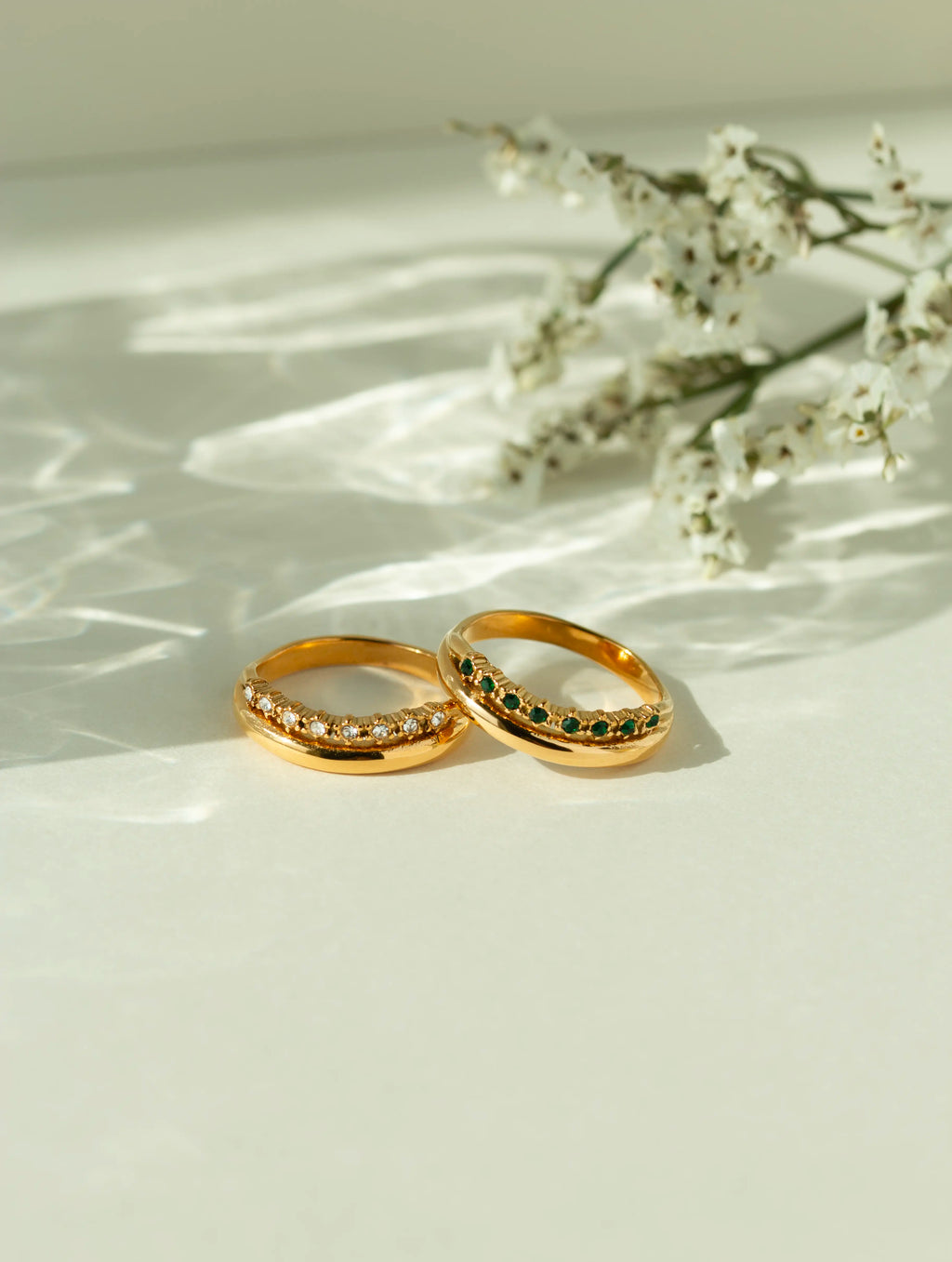 Pair of 18k gold curved gemstone rings featuring a clear diamond band and a green emerald band. Elegant contoured wedding rings or stackable chevron bands shown on a sunlit background.