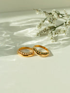 Pair of 18k gold curved gemstone rings featuring a clear diamond band and a green emerald band. Elegant contoured wedding rings or stackable chevron bands shown on a sunlit background.