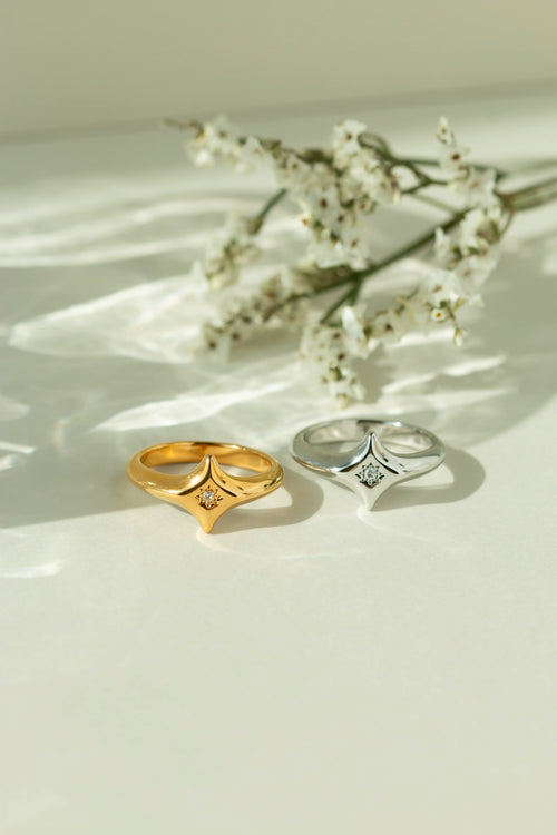 Gold and silver starburst signet rings featuring a central diamond accent stone. Chunky minimalist celestial jewelry styled on a light surface with white flowers.