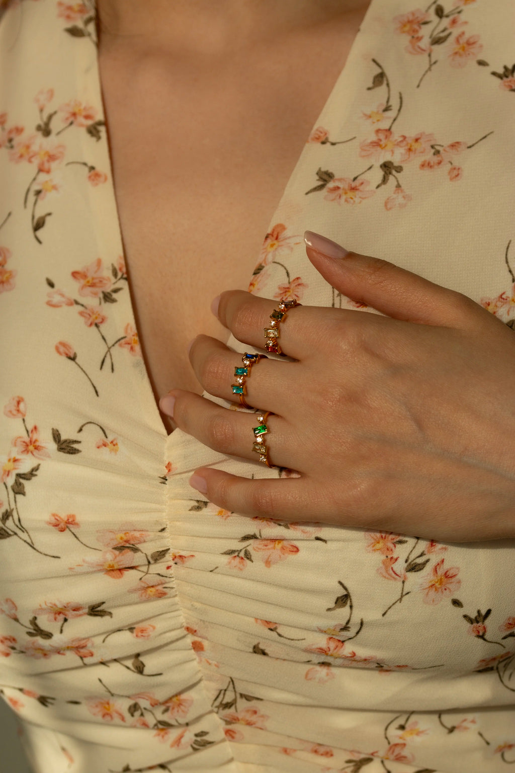 Scattered baguette crystal ring set in yellow gold, unique multi stone family birthstone ring jewelry gift idea for women. Delicate rainbow cluster stacking bands on a cream surface.