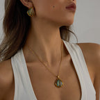 Woman wearing a gold necklace with a pendant and matching earrings against a neutral background