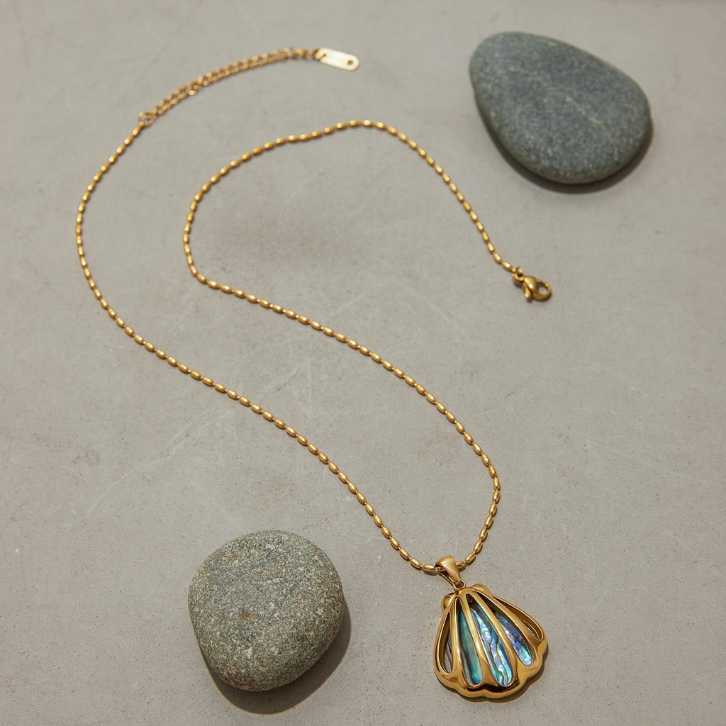 Gold necklace with a shell pendant on a gray surface with stones.