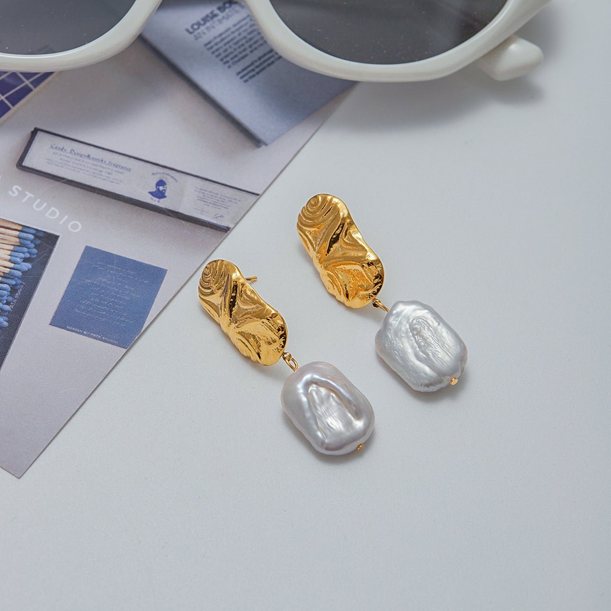 Gold and pearl earrings on a light gray surface with sunglasses in the background.