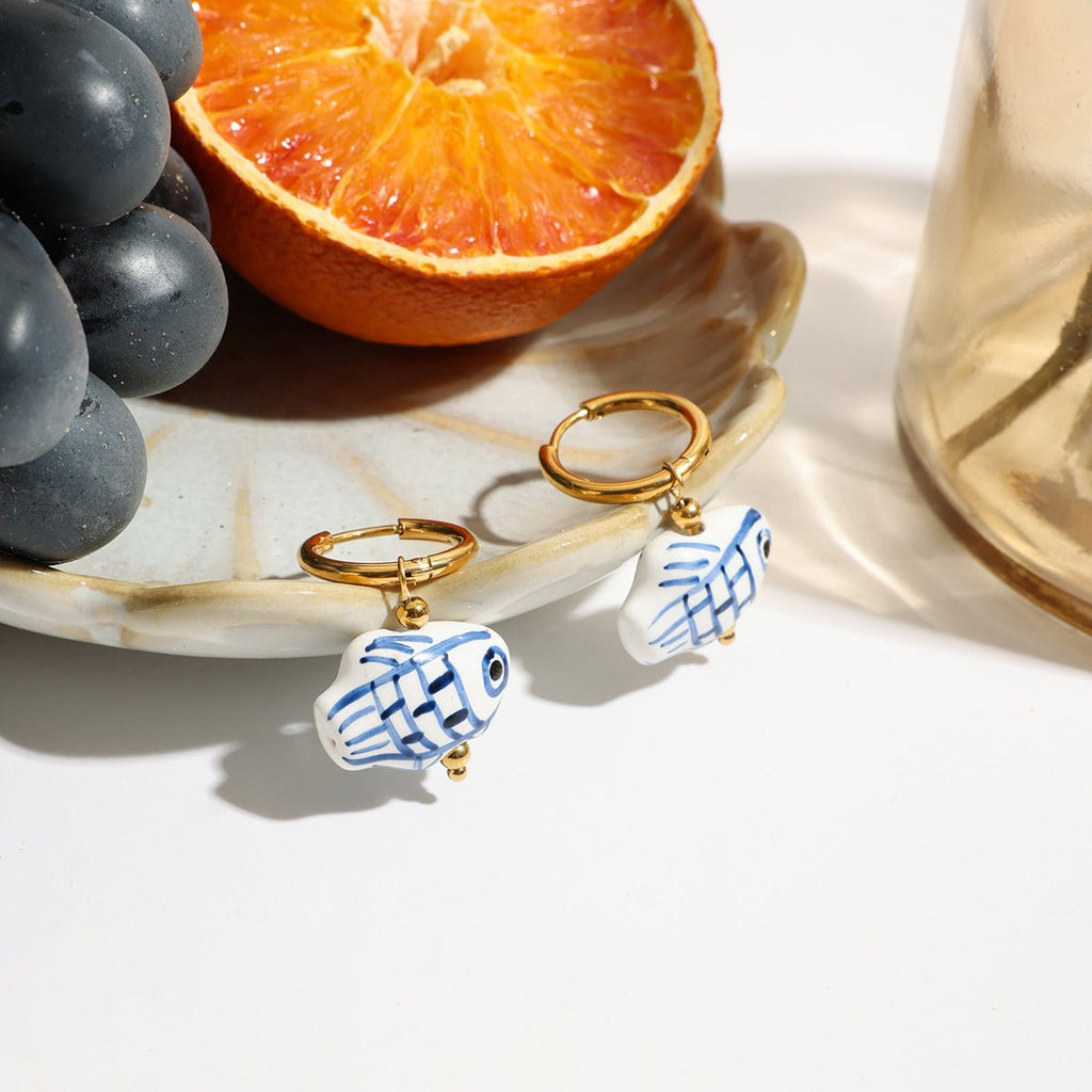 Pair of fish-shaped earrings with gold hooks on a white surface with grapes and an orange.