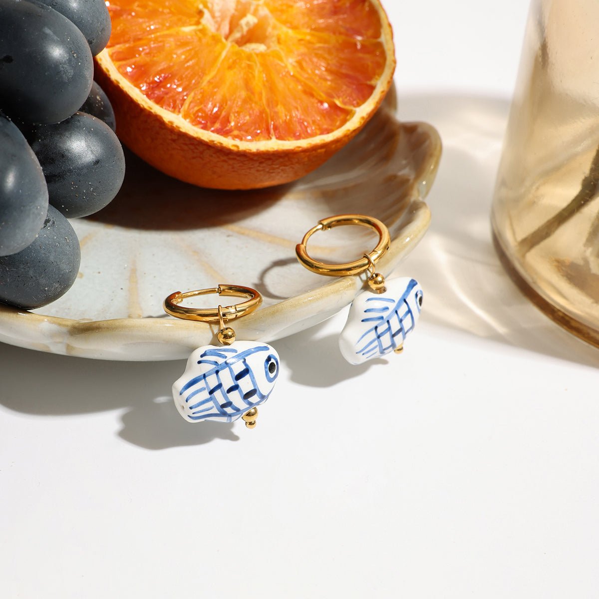 Pair of fish-shaped earrings with gold hooks on a white surface with grapes and an orange.